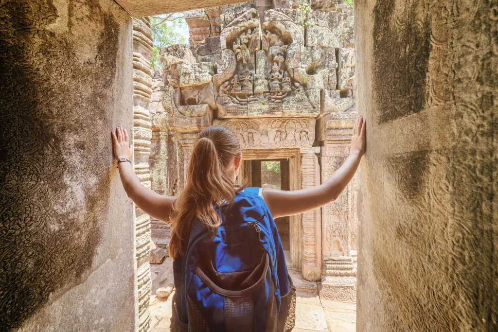 Young female tourist with blue backpack coming to ancient Preah Khan temple in Angkor and looking at the bas-reliefs on the walls and on mysterious ruins. Siem Reap, Cambodia. Toned image.