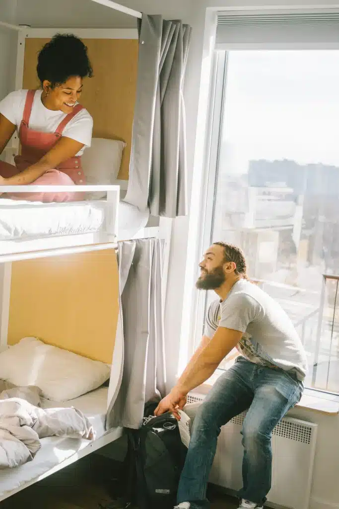 a man packing his backpack next to a bunk bed where a woman lays on the top bunk, smiling down as they talk