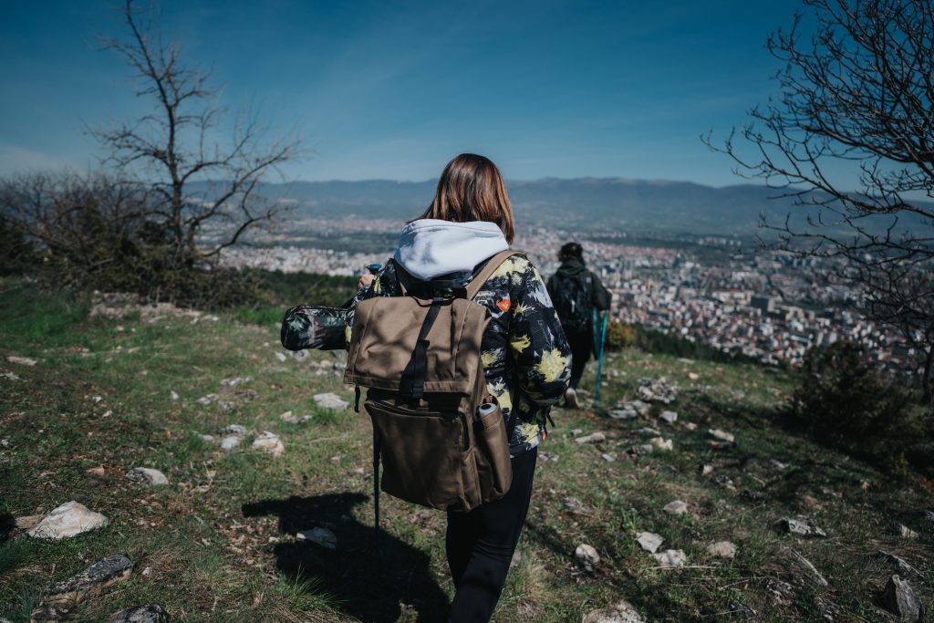 Two individuals hiking downhill on a sunny day overlooking a scenic cityscape from the vantage of a rocky hillside. Individuals with backpacks hike along a grassy trail surrounded by sparse trees, descending towards a city view. 